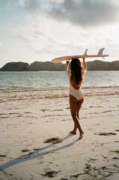 白天，穿着白色比基尼、手持白色冲浪板的女子站在沙滩上